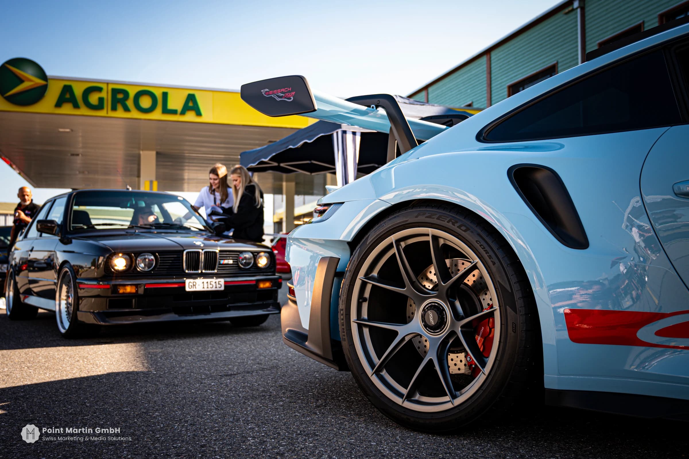 Heckflügel und Rad eines hellblauen Porsche neben einem klassischen schwarzen BMW